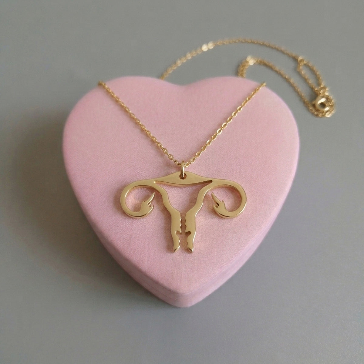 Uterus-Halskette in Gold aus Edelstahl mit Gebärmutter-Anhänger und Mittelfinger auf rosa Herz