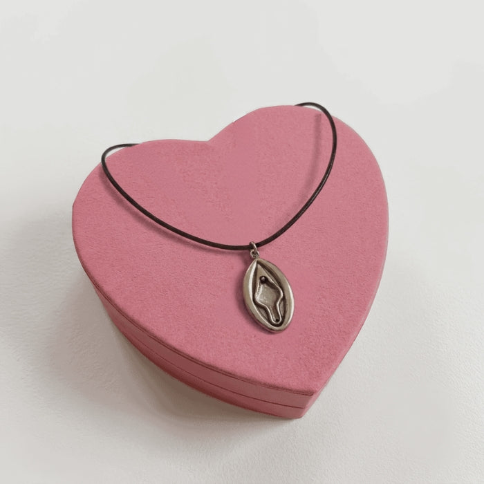 Vulva Kette mit silbernem Vulva-Anhänger aus Metall an schwarzem Band auf rosa Herzschachtel
