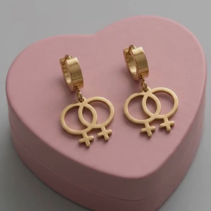 Video: Doppel Venus Symbol Ohrringe in Gold – feministische Edelstahl-Creolen mit zwei verschlungenen Frauensymbolen ♀♀ auf rosa Herzschachtel in Nahaufnahme, Bling Bling Feministas